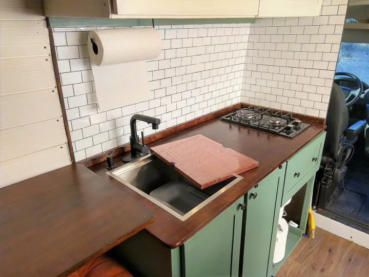 This RV rental kitchen boasts dark counters, white subway tile, sink, stovetop, and green cabinets for comfy travels.. Dodge Promaster 2500 High Roof 2020