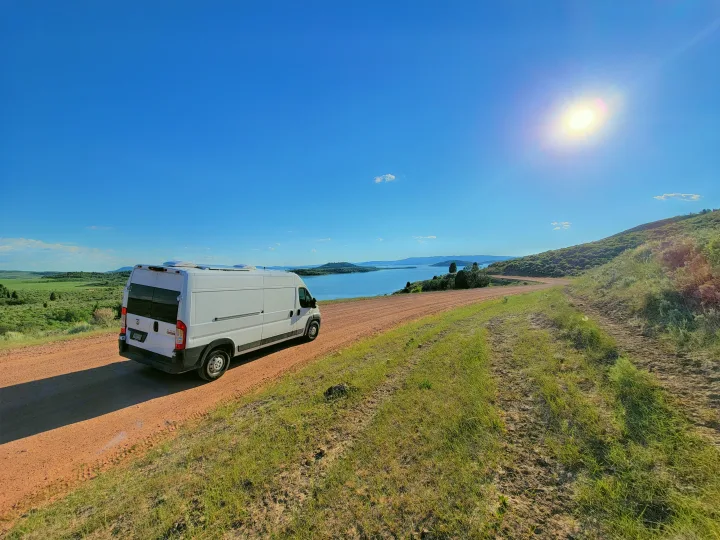White RV rental van on a dirt road, overlooking a vast lake and hills under a sunny sky.. Dodge Promaster 2500 High Roof 2020