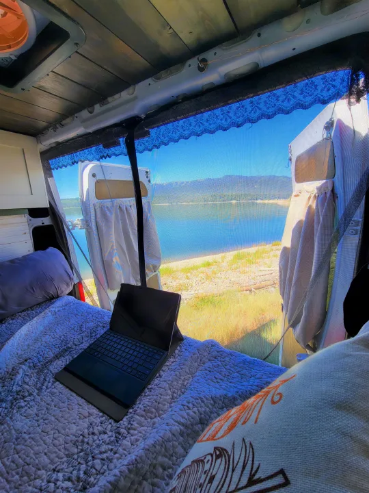 Inside an RV rental: Bed with a laptop, offering a scenic lake and mountain view through screened doors for remote work.. Dodge Promaster 2500 High Roof 2020