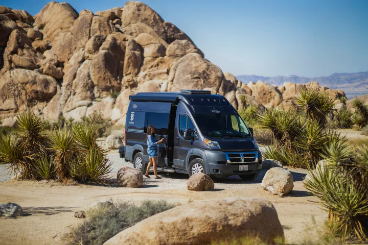 An RV rental camper van parked in a sunny desert landscape with large rock formations. A woman exits the side door.. Hymer Axion Other 2019