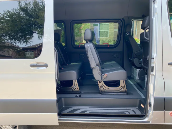 Inside an RV rental van: spacious dark interior with two rows of black passenger seats and open sliding door for easy access.. Mercedes-Benz Sprinter 2021