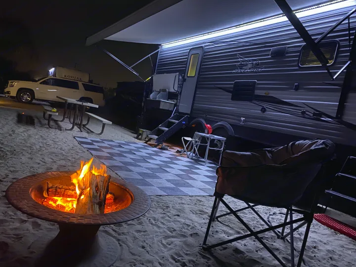 Relaxing night scene with an RV rental. A glowing campfire, illuminated travel trailer, checkered mat, and SUV on sandy terrain.. Jayco Jay Flight 2020