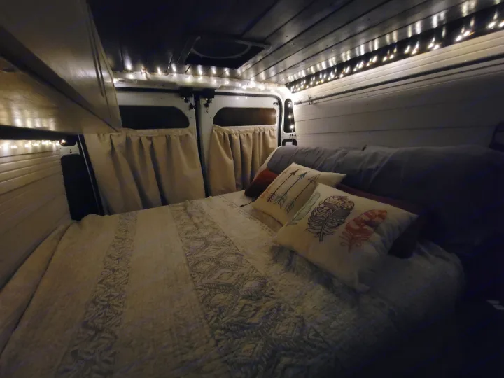 Experience a cozy night in this RV rental! Interior of a camper van bedroom with string lights, quilt, & pillows.. Dodge Promaster 2500 High Roof 2020