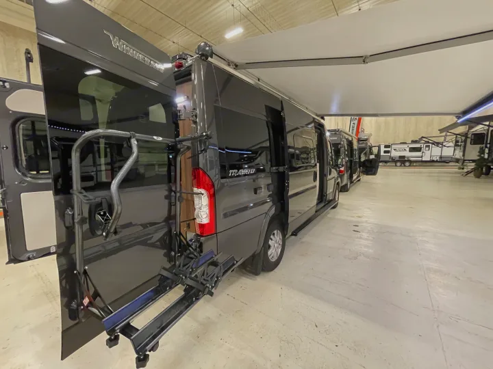 Gray Winnebago Travato camper vans with open doors, one with a bike rack, on display in an RV rental showroom.. Winnebago Travato 2022