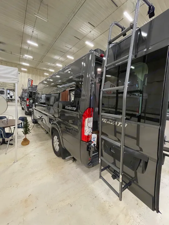 Dark grey Travato camper van, shown on its side, parked indoors. This RV rental unit features a rear ladder and tail lights.. Winnebago Travato 2022
