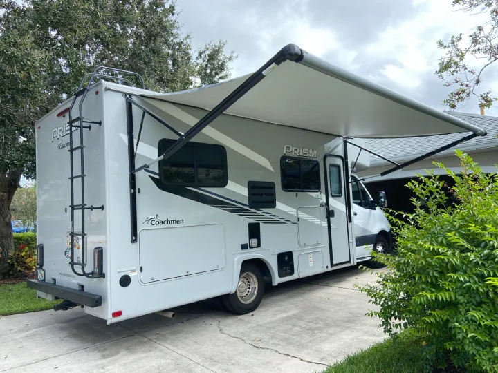 A white and gray Class C motorhome with an extended awning is parked on a concrete driveway, ready for an RV rental adventure.. Mercedes-Benz Sprinter Coachmen Prism 2021