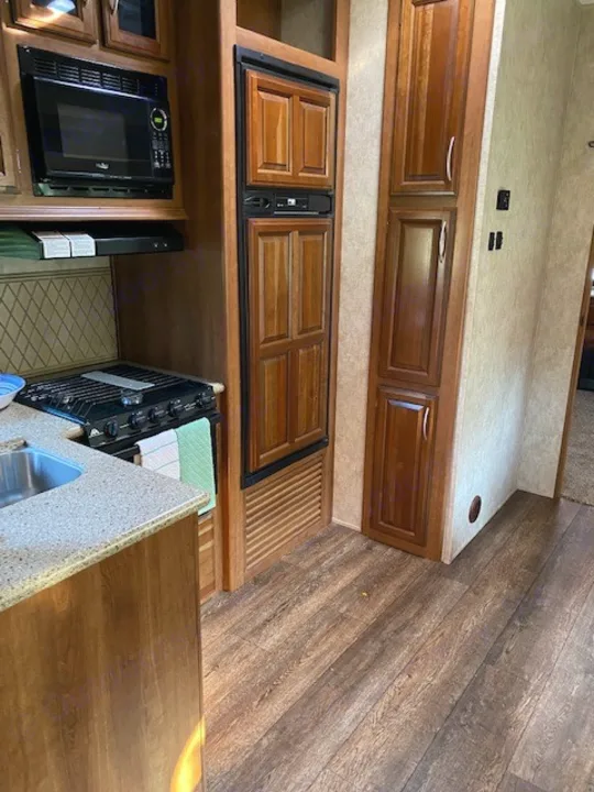 Bright RV rental kitchen featuring a microwave, stove, wood-paneled fridge, and brown storage cabinets.. Forest River Crusader BH 2015