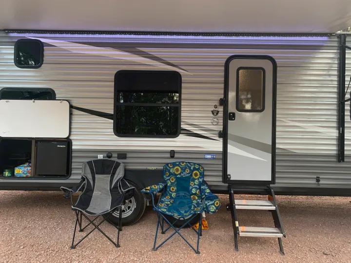 RV rental campsite setup: extended awning with lights, accessible outdoor fridge, chairs, and entry steps on gravel.. Jayco Flight 2021