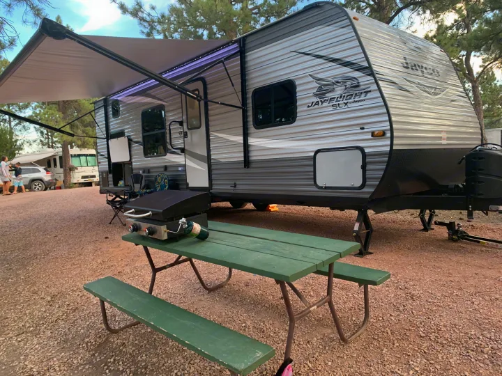 A gray travel trailer RV rental at a scenic campsite. Features include an extended, lit awning, picnic table, and grill setup.. Jayco Flight 2021