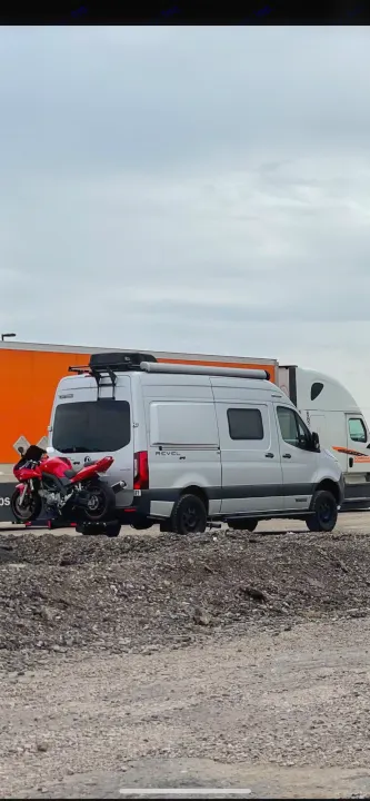 Adventure-ready RV rental: Silver Winnebago Revel camper van with a red motorcycle on a hitch carrier. Parked near semi-trucks.. Winnebago Revel 2021
