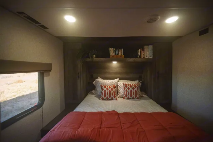 Inside this cozy RV rental bedroom: a bed with coral bedding, decorative pillows, overhead storage, and a desert view.. KZ Sportsmen 2021