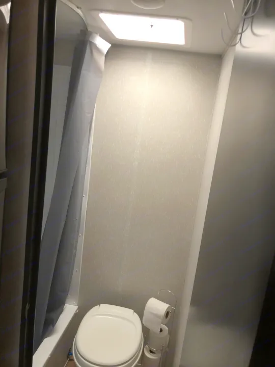 Compact RV rental bathroom with a white toilet, gray shower curtain, ceiling light, and a chrome toilet paper holder.. Forest River Wolf Pup 2021