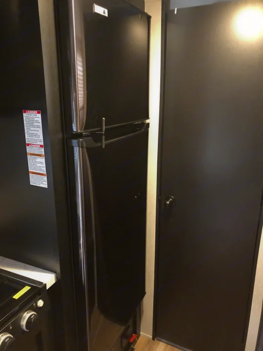 Inside an RV rental, this view shows a black RV refrigerator with safety labels, a stovetop, and an interior door.. Forest River Wolf Pup 2021
