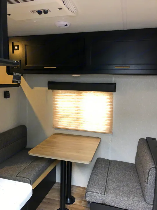 Cozy RV rental dinette featuring gray upholstered benches, light wood table, black overhead cabinets, and window.. Forest River Wolf Pup 2021