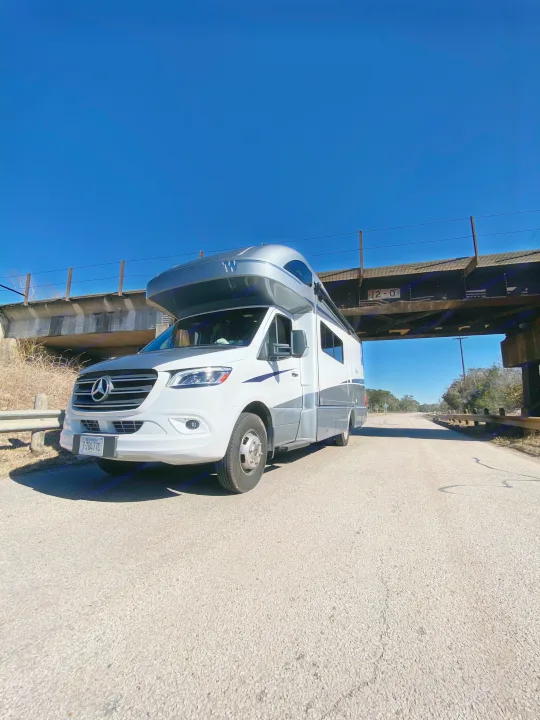 White and grey RV rental, a motorhome, parked on an asphalt road under a railway bridge on a clear day.. Winnebago View 2022