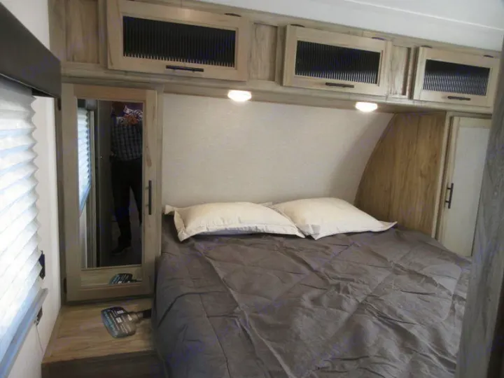Cozy RV rental bedroom with a gray bed, overhead cabinets, and mirrored closet reflecting a person, ideal for travel.. Forest River 30DBH-L 2021