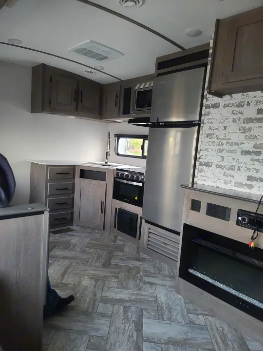 RV rental interior: modern kitchen with stainless steel fridge, oven, microwave, light wood cabinets, and brick accent wall.. Keystone Springdale 2021