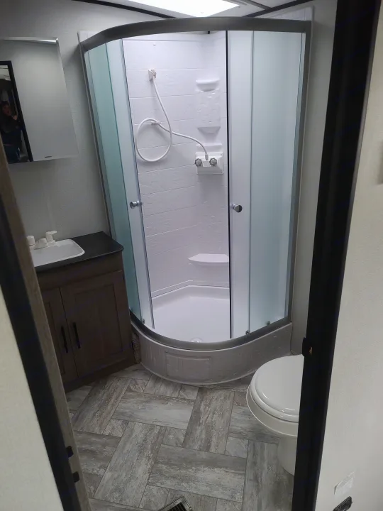 Compact RV rental bathroom featuring a glass shower, white toilet, dark vanity, and stylish gray tile flooring.. Keystone Springdale 2021