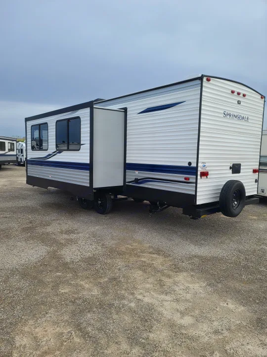 White & blue Springdale travel trailer, ideal for RV rental, parked on gravel. Features include a rear spare tire and extended slide-out.. Keystone Springdale 2021