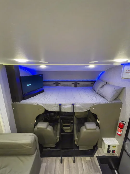 Cozy RV rental cab-over bunk featuring a bed, TV, and driver's cockpit below, illuminated by blue LED lights.. Jayco Redhawk 2021