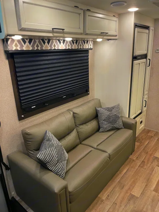 Modern RV rental interior with a gray sofa bed, patterned window valance, upper cabinets, and a refrigerator.. Jayco Redhawk 2021