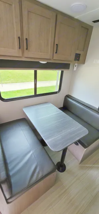 RV rental interior: A compact dinette with dark benches, light table, wood cabinets, and a window view of a path.. Keystone Hideout 2021