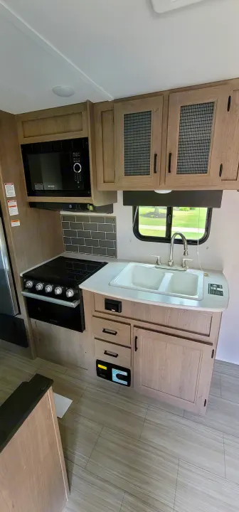 Modern RV rental kitchen featuring light wood cabinets, sink, microwave, stove, and a window with green views.. Keystone Hideout 2021