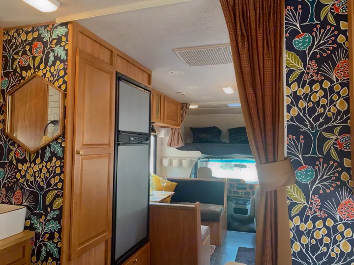 Interior of an RV rental showing custom botanical wallpaper, wood cabinets, fridge, and a cozy living area with bunk.. Winnebago Chalet 2010