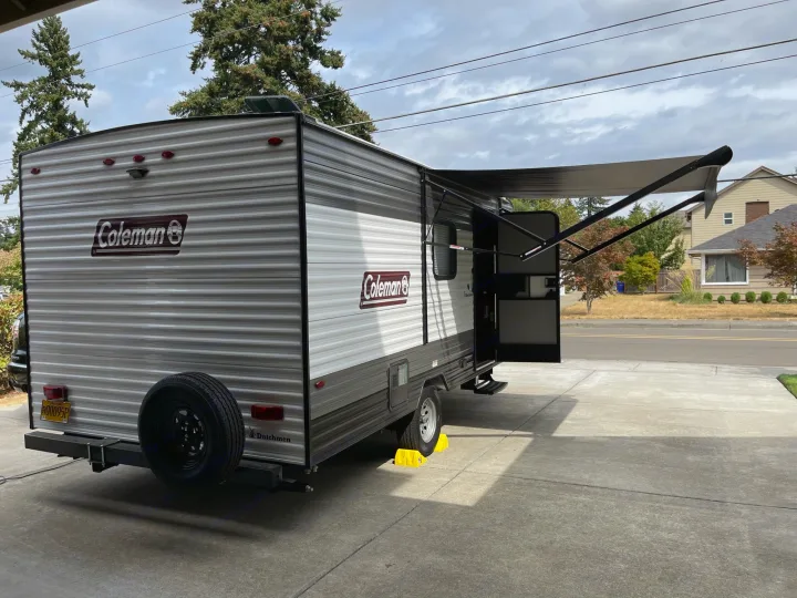 This grey Coleman travel trailer, ready for RV rental, is parked with awning extended & a rear spare tire. Wheel chocks visible.. Other Other 2021