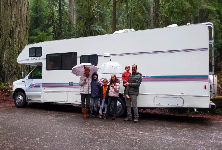 Family trip by RV rental: Parents and four kids with umbrellas in a redwood forest with their Itasca RV.. Itasca Spirit 1995