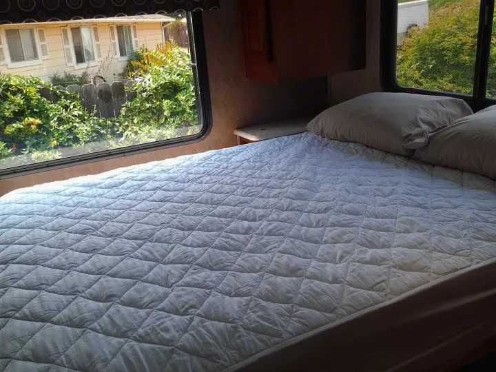 Comfortable RV rental bedroom with a quilted bed, pillows, and bright windows offering green outdoor views. Your cozy home away from home.. Forest River Sunseeker 2018