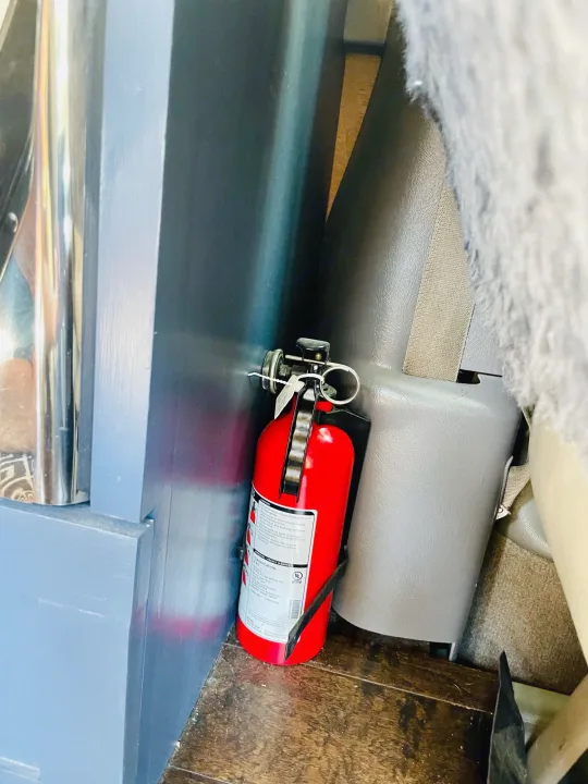 A red fire extinguisher, crucial for RV rental safety, is mounted on the floor next to interior paneling and a seatbelt.. Chevrolet Astro Van AWD 2003