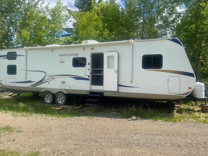 A white "North Country Lakeside" travel trailer (RV rental) with its door open and steps down, parked on gravel near green trees.. Heartland North Country Lakeside 2011