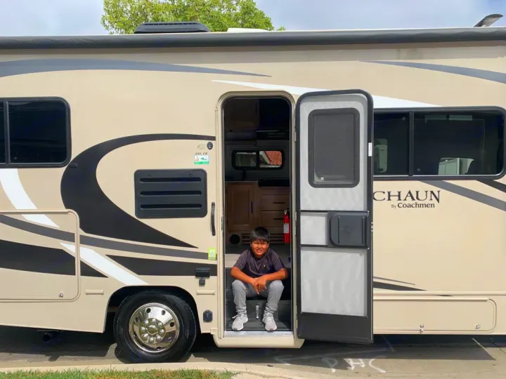 A young boy smiles from the open doorway of a beige Class C RV rental, a Coachmen motorhome. Ready for travel!. Coachmen Leprechaun 2021