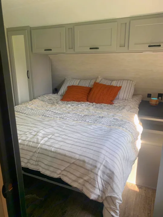 Compact RV rental bedroom featuring a striped bed with orange pillows, overhead cabinets, and a side wardrobe.. Forest River Vibe 2021