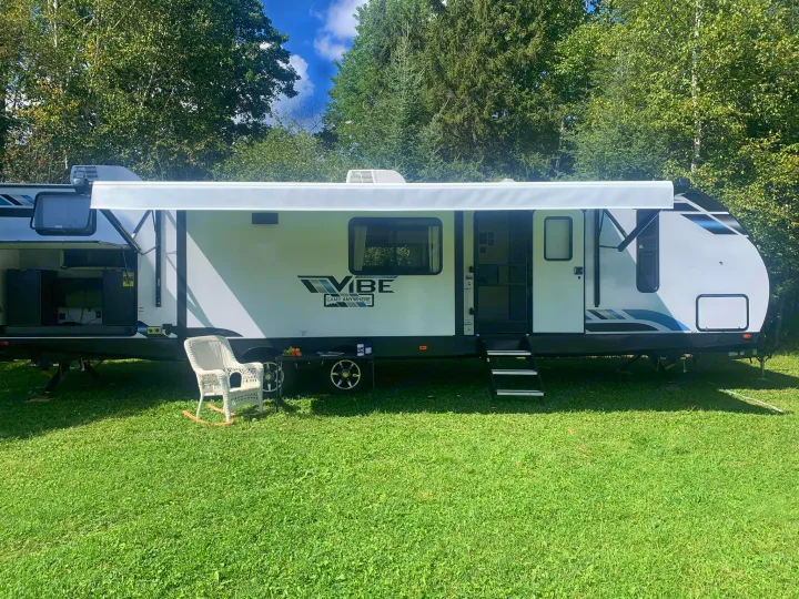 White RV rental with extended awning, wicker chair, and steps on green grass at a sunny campsite under trees.. Forest River Vibe 2021