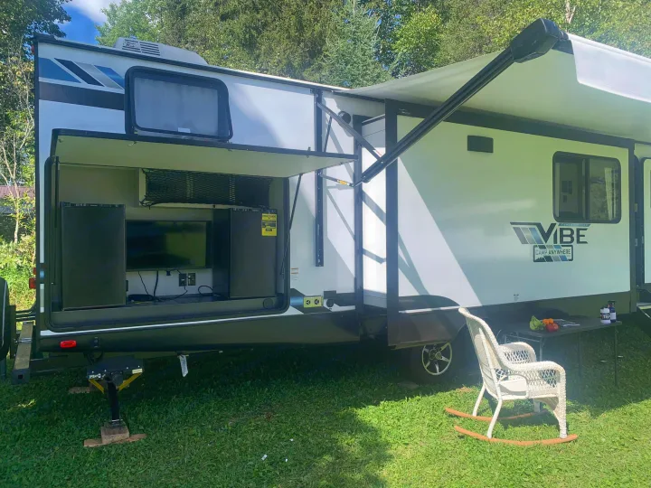 RV rental: A white camper's outdoor entertainment area with TV, awning, and a wicker chair on grass.. Forest River Vibe 2021