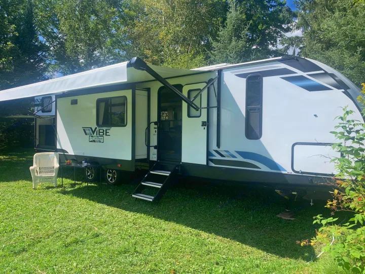 Relax in nature with this RV rental! A white Vibe travel trailer, awning extended, steps down, on a sunny green campsite.. Forest River Vibe 2021