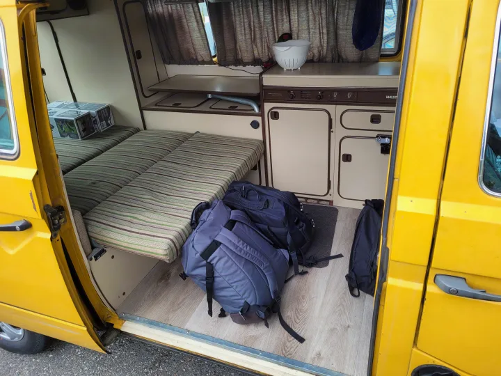 Inside a yellow RV rental, showing striped beds, a kitchenette with sink, and backpacks on wood-look flooring.. Volkswagen Westfalia 1982