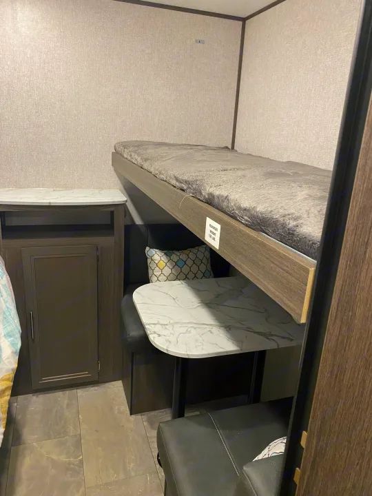 Inside this RV rental: a cozy bunk bed (150 lbs max), a small dinette with a marble-look table, and built-in storage. A compact space for travel.. Dutchmen Kodiak 2021