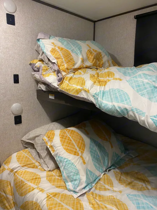 An RV rental's cozy bunk beds, dressed in vibrant yellow and blue leaf-patterned bedding, are equipped with wall lights and USB chargers.. Dutchmen Kodiak 2021
