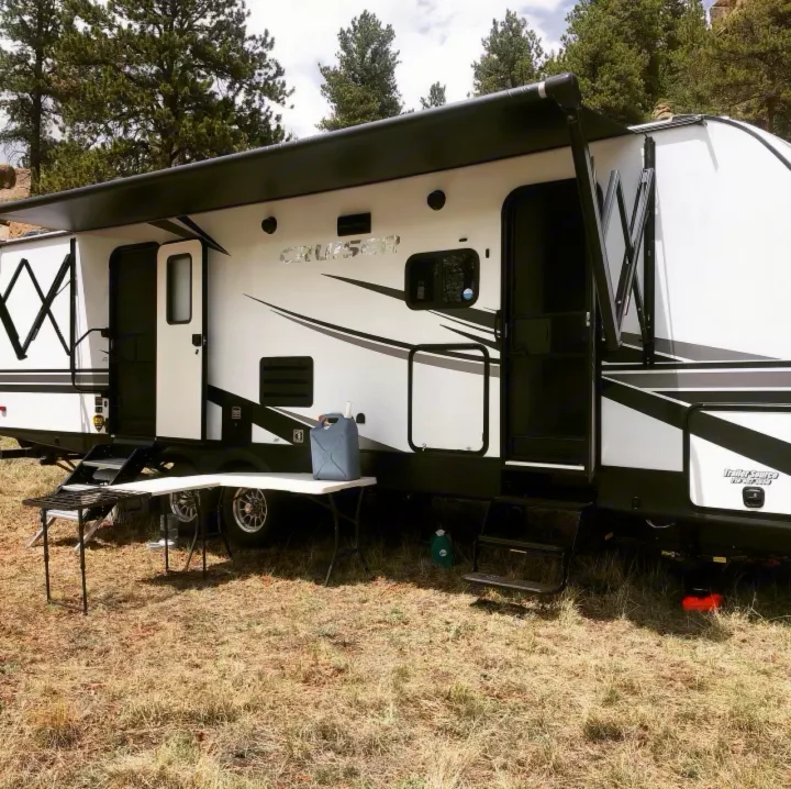 An RV rental, a white and black Cruiser travel trailer, is set up for camping in a grassy area amidst pine trees.. Crossroads Cruiser Aire 2021