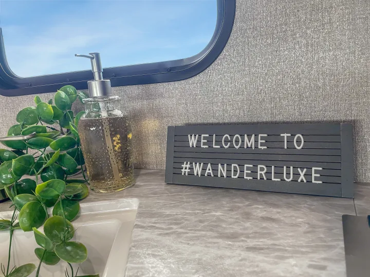 RV rental interior with a "Welcome to #Wanderluxe" sign, soap dispenser, and plant. Blue sky visible through window.. Jayco Jay Flight 2021