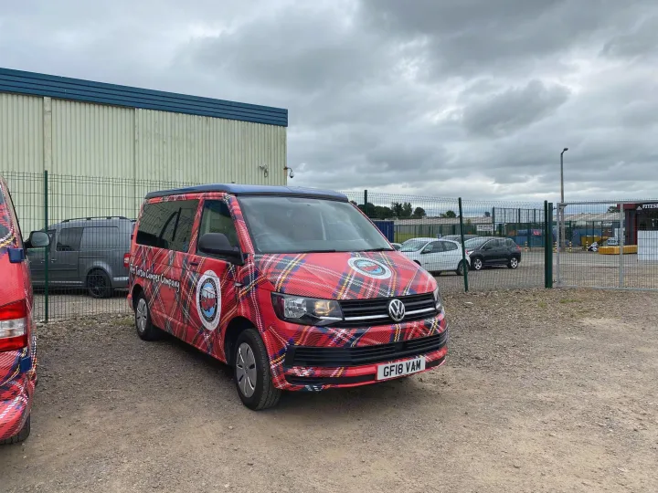 A vibrant red tartan VW campervan, branded "The Tartan Camper Company," parked on a gravel lot. A unique choice for rv rental.. Volkswagen T6 2018