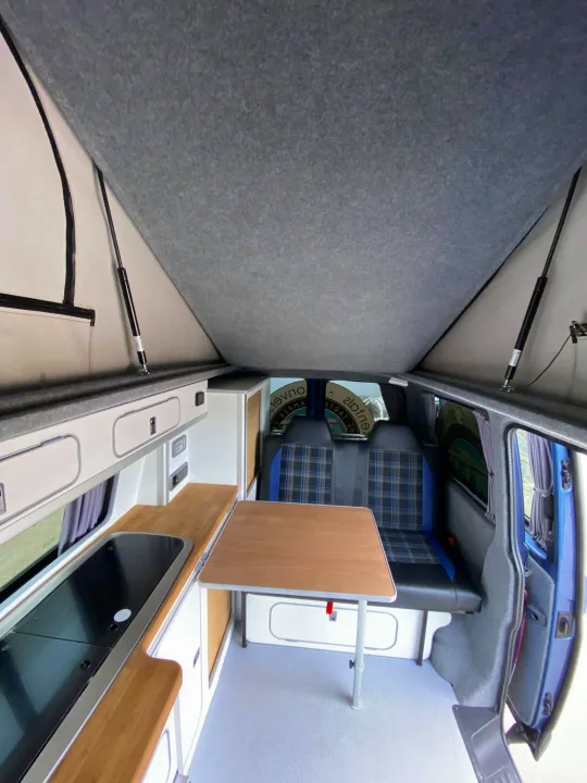 This spacious camper van interior, ideal for **rv rental**, features a pop-top roof, kitchenette, table, and plaid seats.. Volkswagen T5 2018
