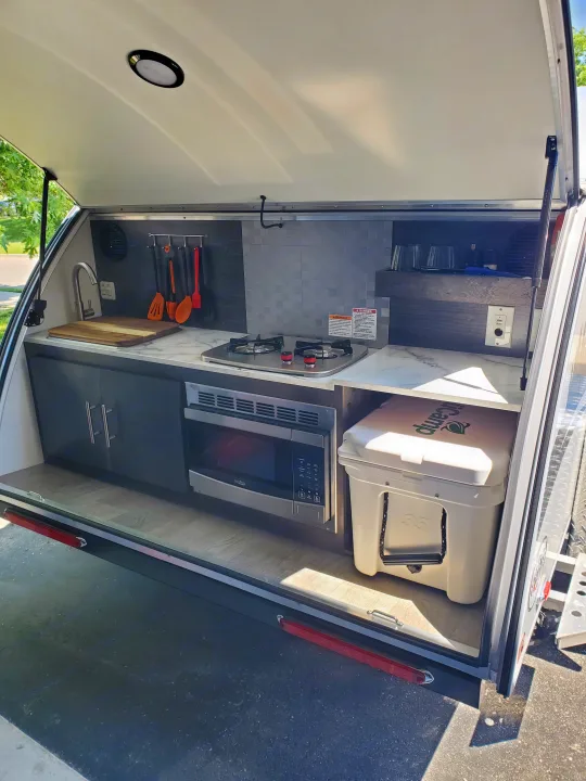 Explore this RV rental's clever outdoor kitchen galley, featuring a sink, stove, microwave, and cooler for camping adventures.. nuCamp T@G 2021
