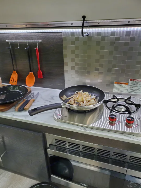 RV rental kitchen with onions cooking on a Suburban stovetop, under-cabinet lighting, and a High Pointe microwave.. nuCamp T@G 2021