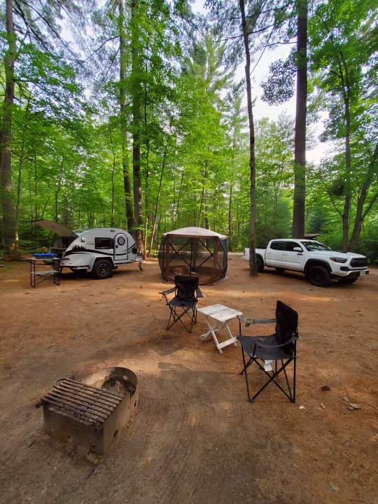 Wooded campsite features a cozy teardrop RV rental, a white truck, a screened tent with a dog, chairs, and a fire pit.. nuCamp T@G 2021