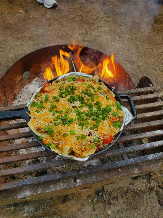 A cast iron skillet of cheesy nachos cooking on a grate over a campfire, ideal for an RV rental trip.. nuCamp T@G 2021