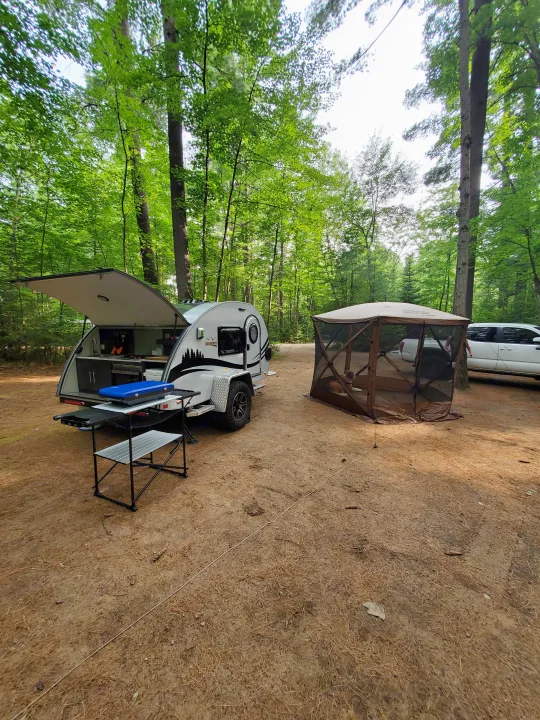 Camping with an rv rental: a teardrop camper with open kitchenette, screen tent, and dog at a wooded campsite.. nuCamp T@G 2021